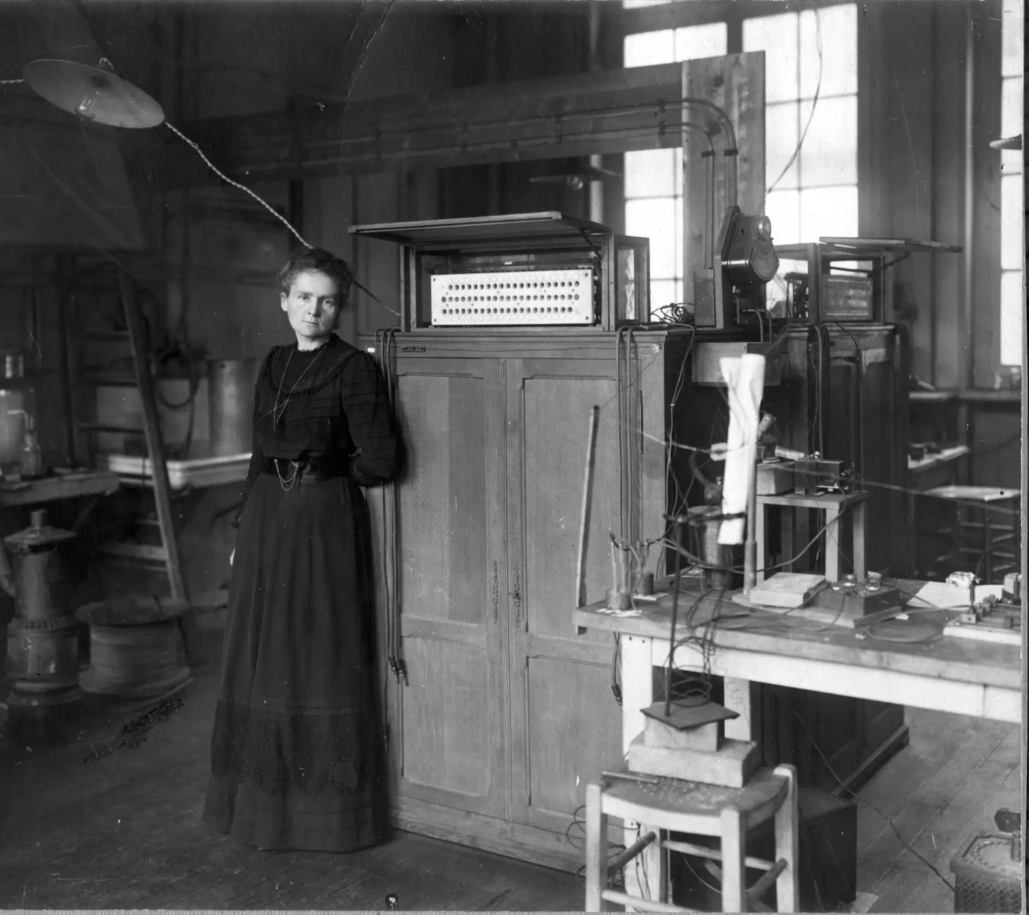 Marie Curie standing beside laboratory cabinets and scientific equipment.