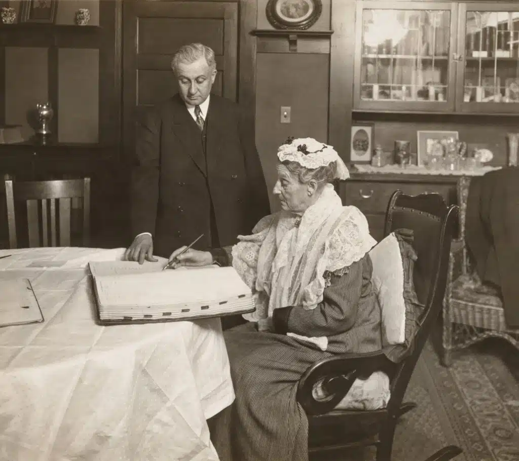 Abigail Scott Duniway seated signing a voter registration book, with County Clerk John B. Coffey standing beside her.