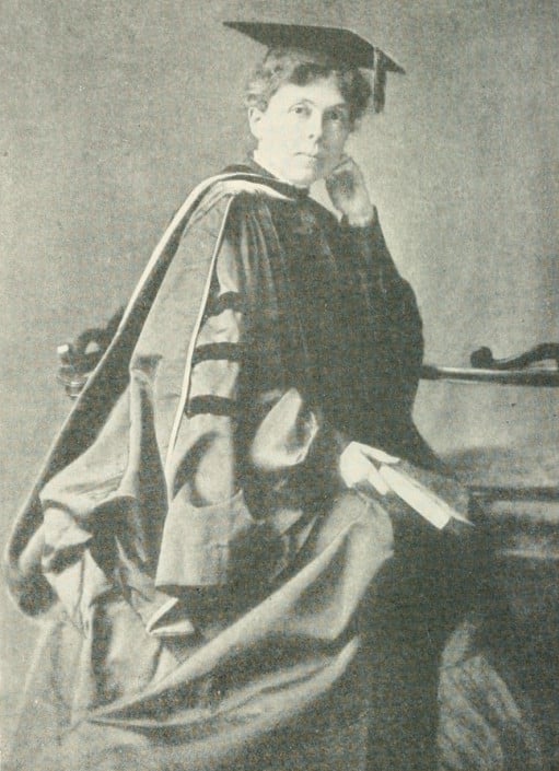 Alice Freeman Palmer wearing academic robes and standing beside a table.