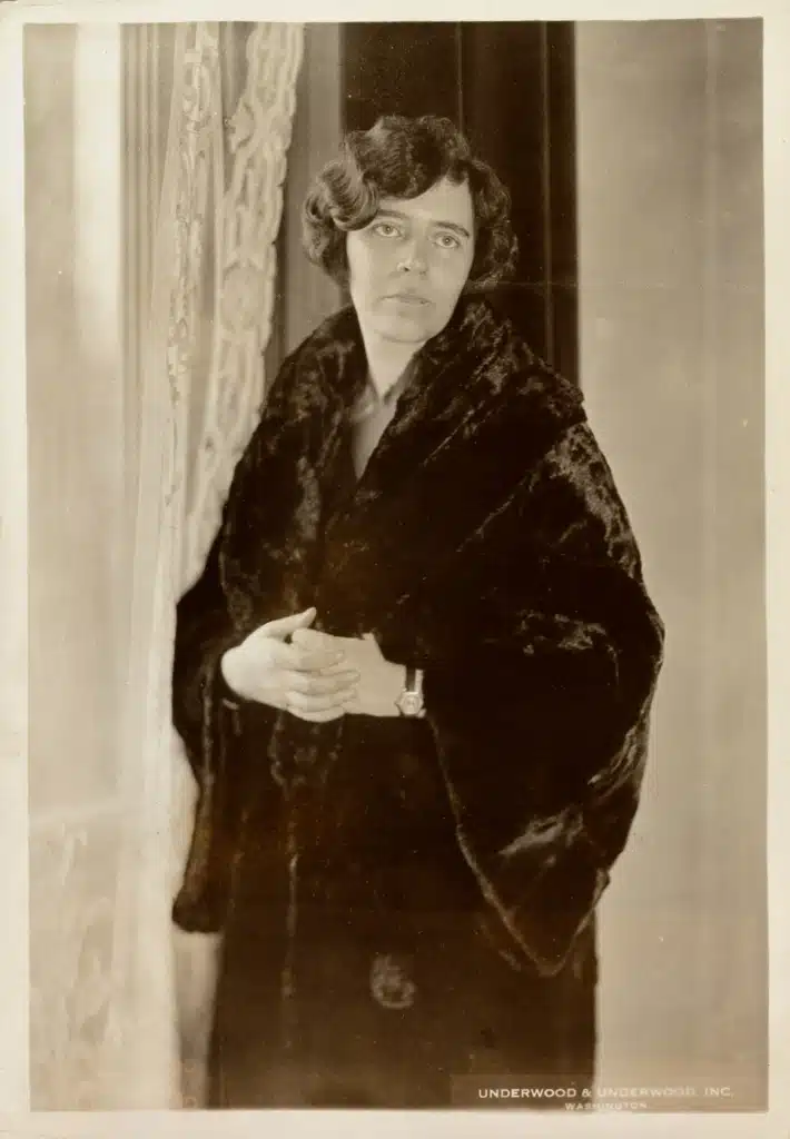 Alice Paul standing beside a curtain in a dark coat.
