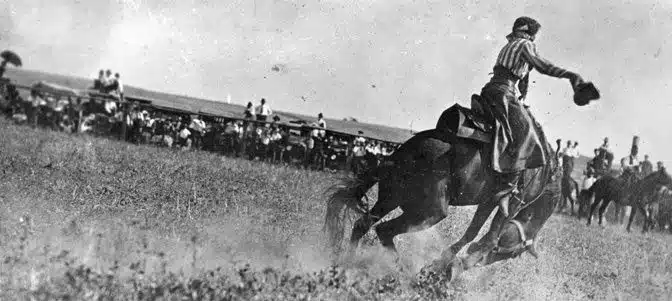 Fannie Sperry Steele rides a bucking horse with spectators in the background.
