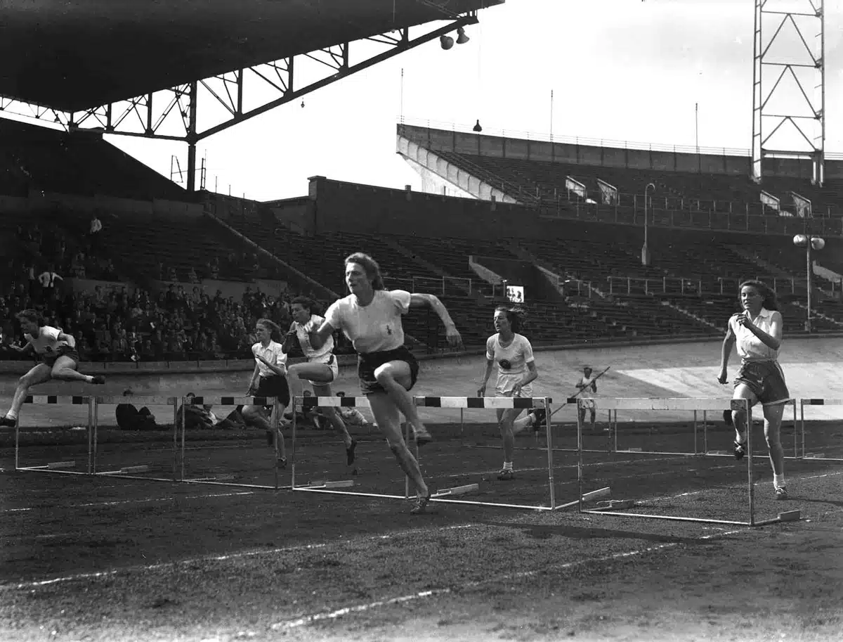 Fanny Blankers-Koen clears a hurdle on a stadium track with other runners nearby.