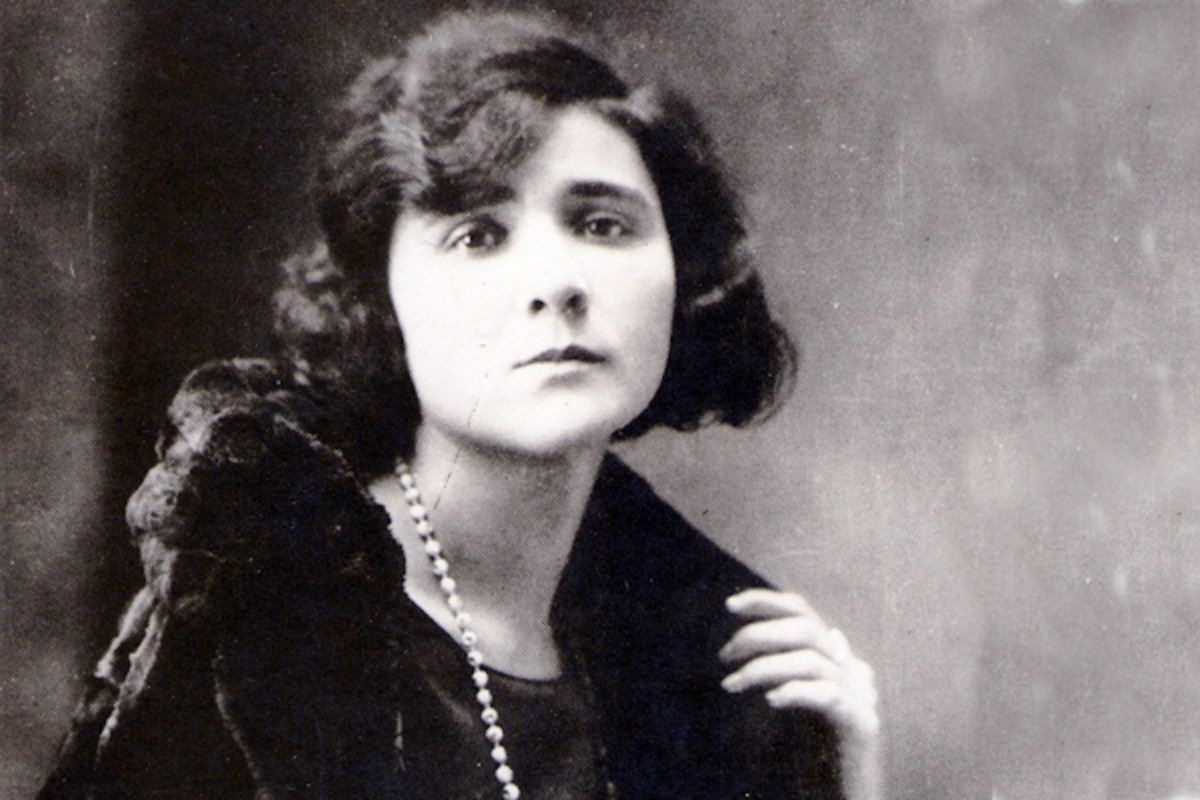 Florbela Espanca wearing a dark fur-collared coat and a long beaded necklace, with one hand raised near her shoulder.