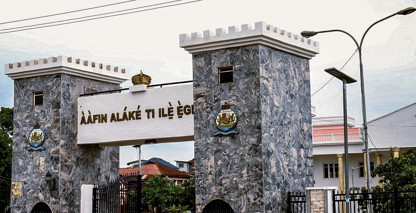 Ake Palace entrance in 2023 | Amazing Women In History Modern entrance gates to Ake Palace, with stone-faced pillars, a white arch and surrounding buildings.