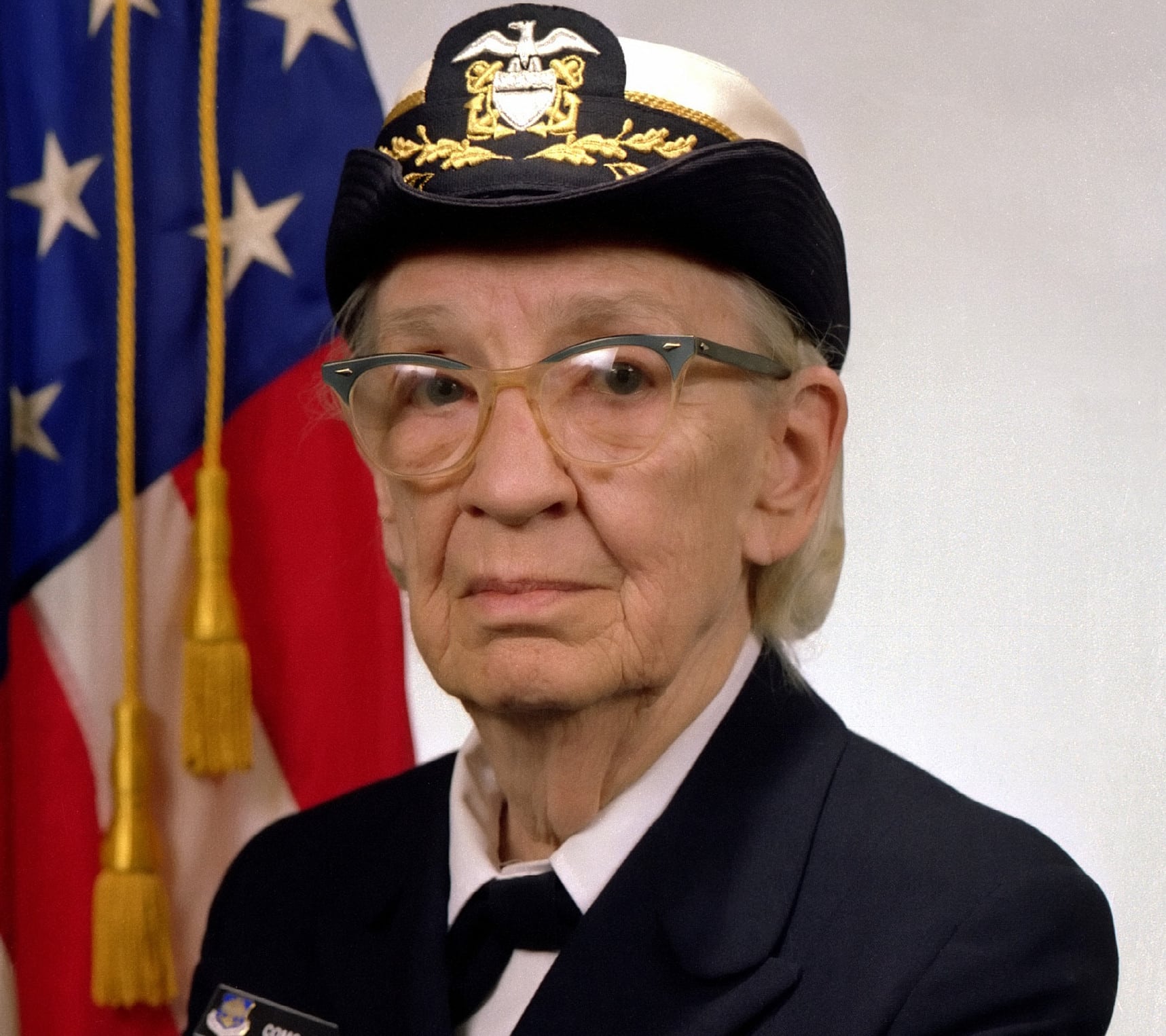 Grace Hopper wearing a U.S. Navy uniform and cap, with an American flag behind her.
