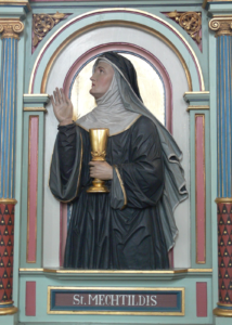 Painted carving of Mechthild of Magdeburg holding a gold chalice and raising one hand, framed by blue and gold columns.