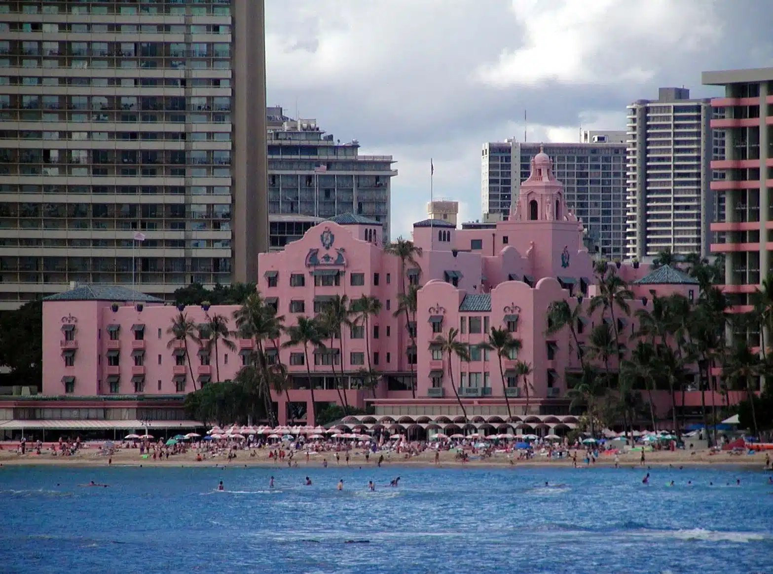 The pink Royal Hawaiian Hotel viewed from the sea, with beachfront buildings around it.