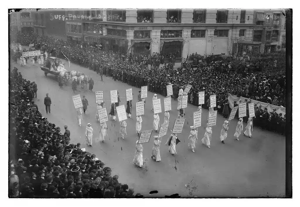 A suffrage parade with banners moves through a crowded New York street.