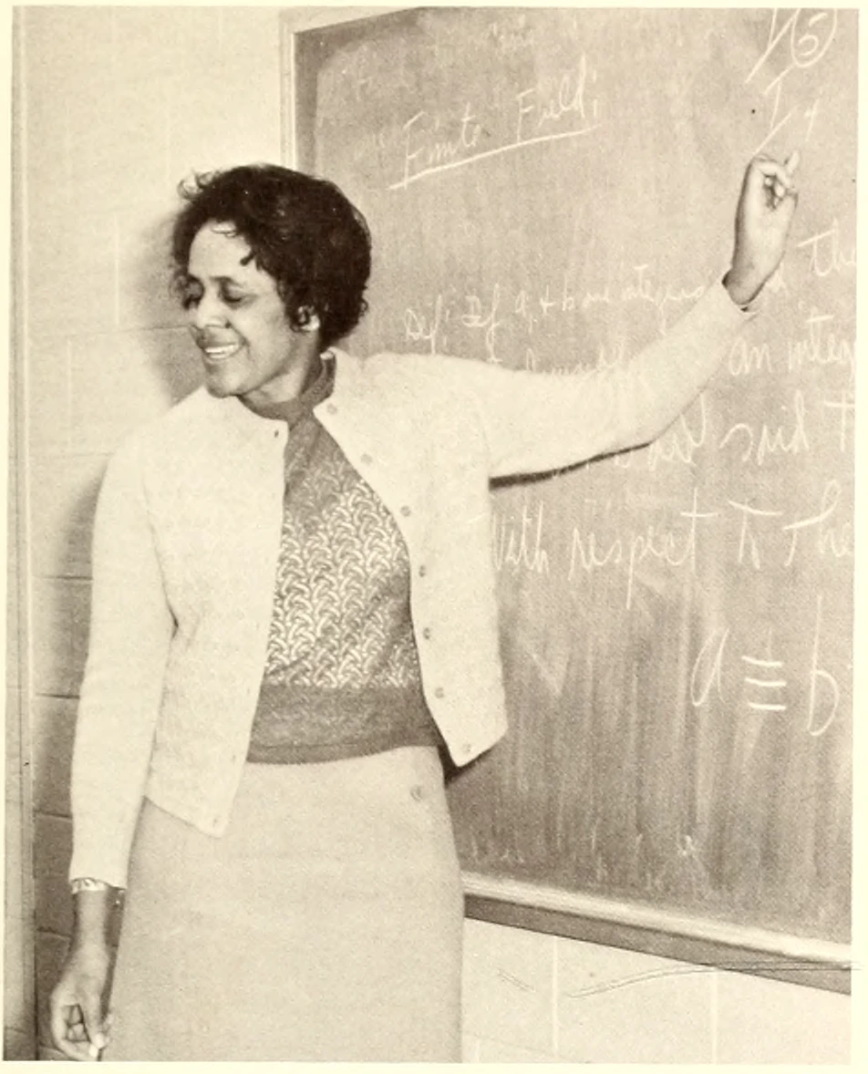 Virginia K. Newell stands at a blackboard, pointing toward a mathematical expression.