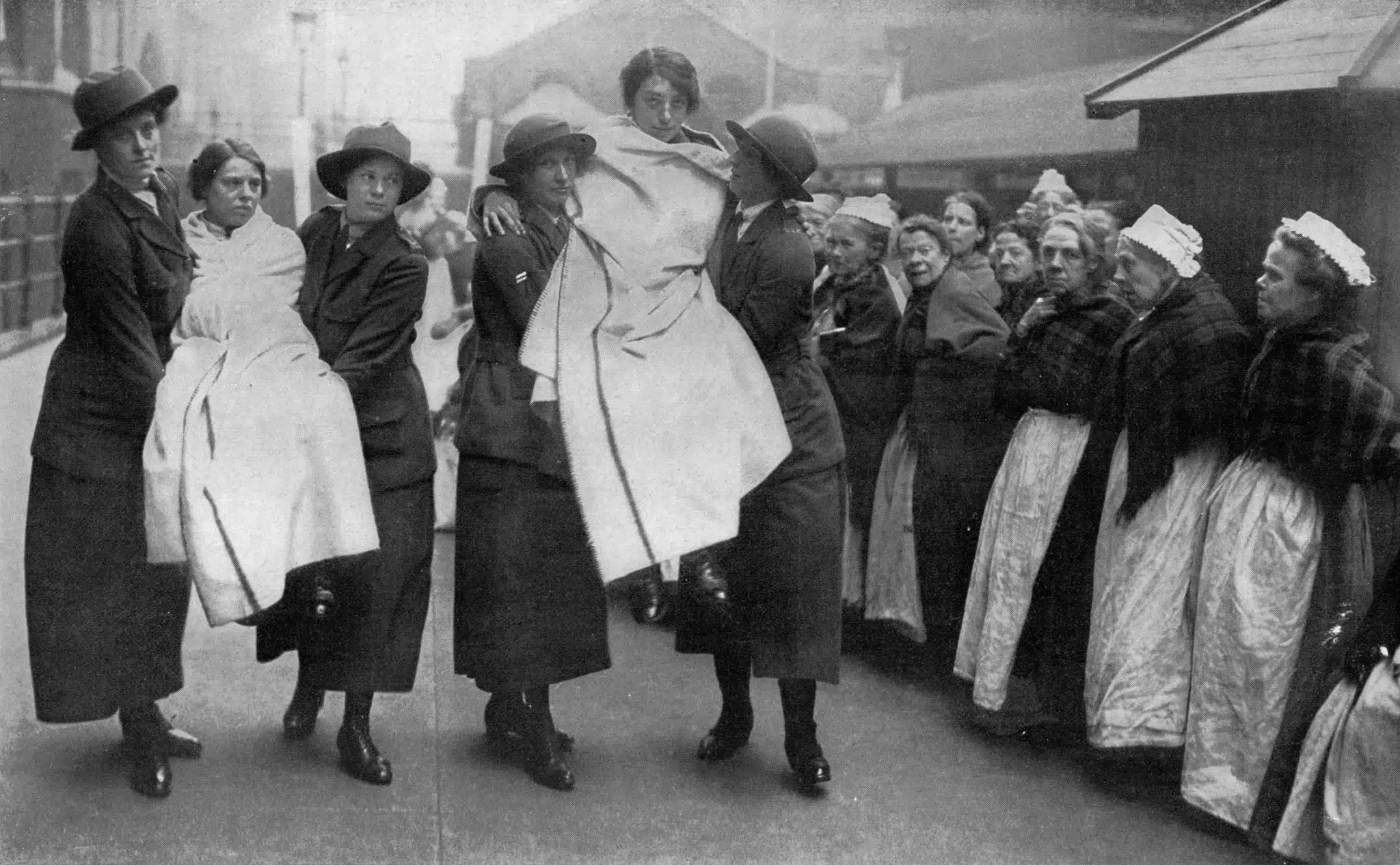 Women demonstrate firefighting equipment as a group watches outside a workhouse.