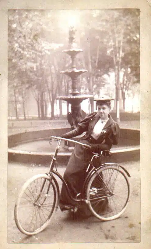 A woman in period clothing poses with a bicycle outdoors.